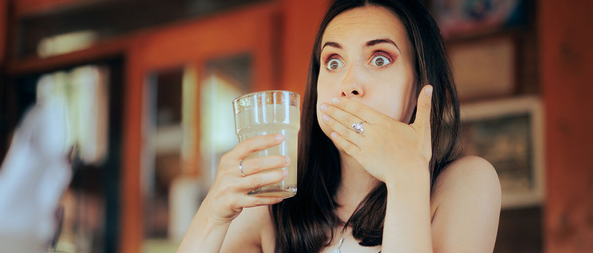 Eine Frau hält ein Glas Limonade in der rechten Hand. Sie schaut erschrocken, hat die Wangen aufgeplustert und hält sich die linke Hand vor den Mund.