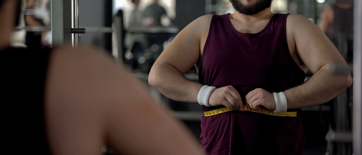 Übergewichtiger Mann nimmt vor dem Spiegel im Fitnessstudio die Maße seines Bauchs