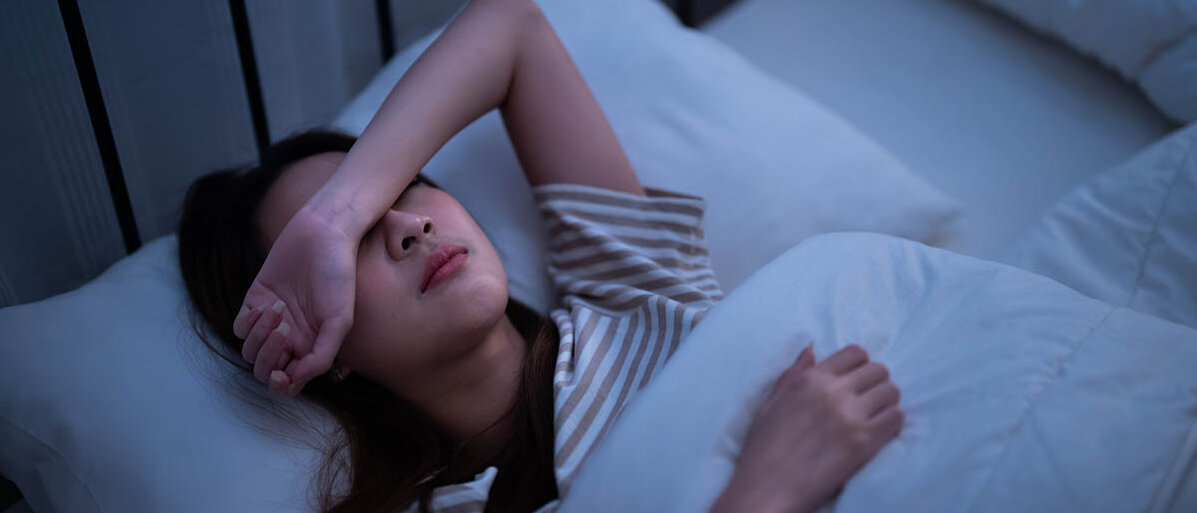 Migräneprophylaxe und Migränetherapie Eine junge Frau liegt im dunklen Zimmer im Bett und hat den Arm über die Augen gelegt.