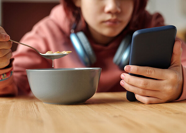 Junges Mädchen mit umgehängten Kopfhörern sitzt am Tisch und hält ihr Handy mit einer Hand fest. In der anderen Hand hält sie einen Löffel für ihr Frühstück.