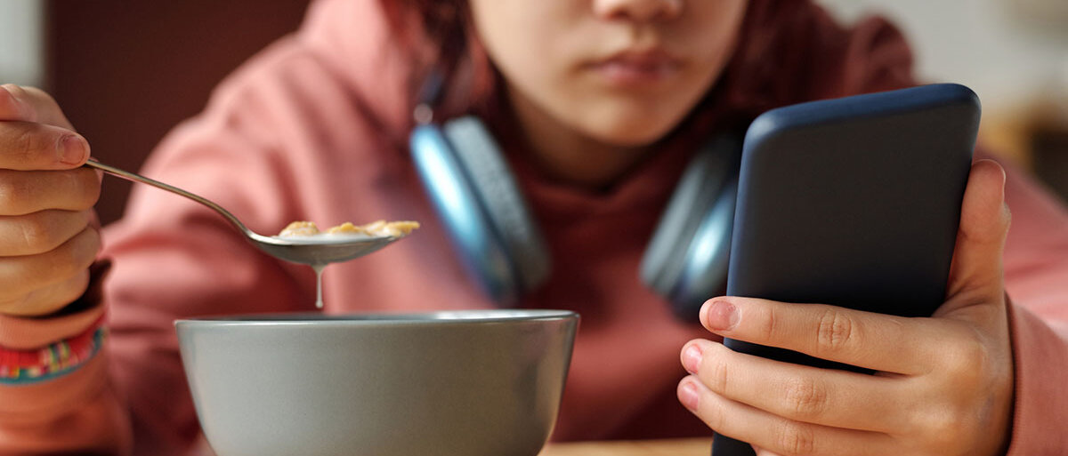 Junges Mädchen mit umgehängten Kopfhörern sitzt am Tisch und hält ihr Handy mit einer Hand fest. In der anderen Hand hält sie einen Löffel für ihr Frühstück.