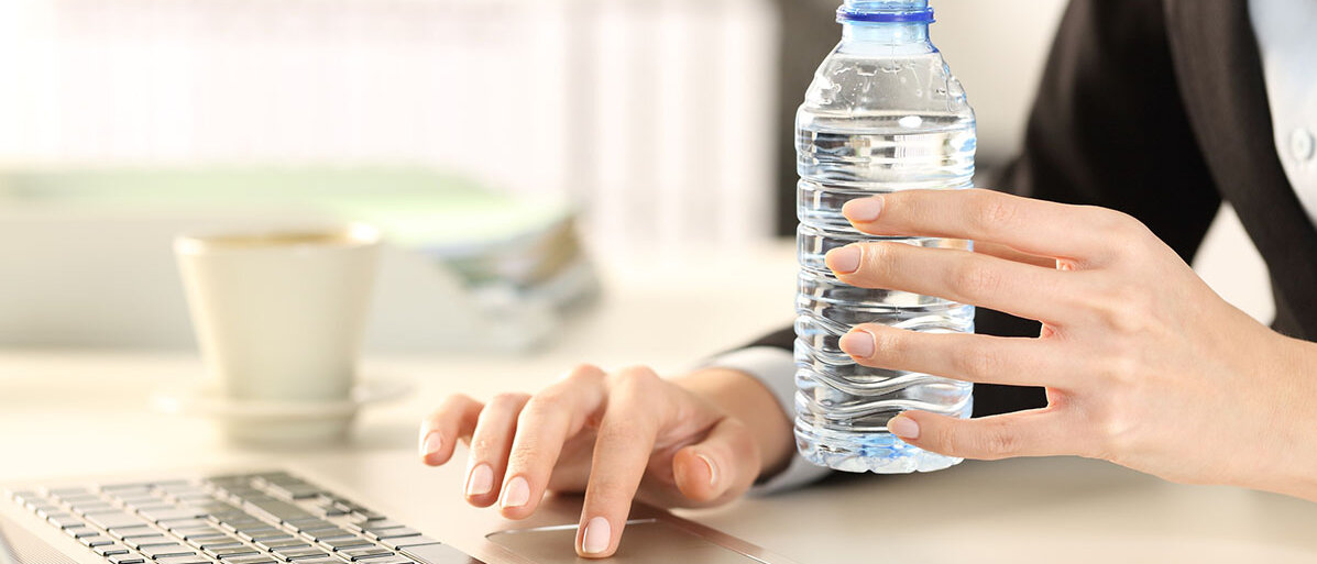 Frau haelt eine Plastikwasserflasche in der Hand am Arbeitsplatz. Vor ihr steht dder aufgeklappte Laptop