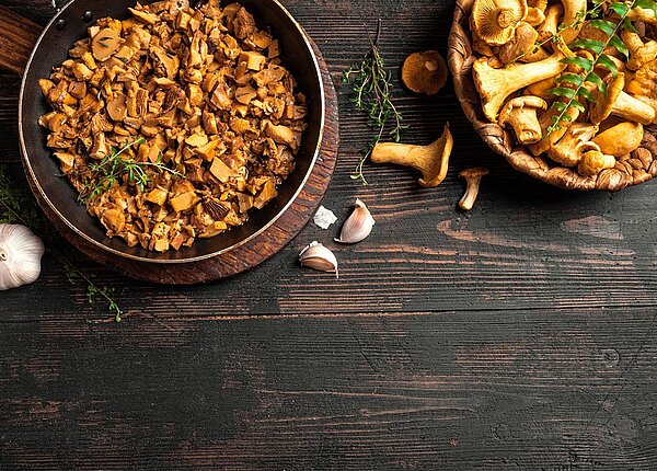 Eine Pfanne und ein Körbchen mit frischen Pfifferlingen auf einem dunklen Holztisch 