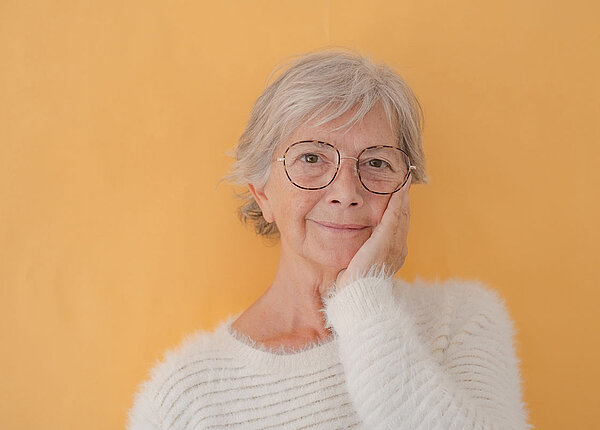 Eine Seniorin mit kurzem grauem Haar hat ihre linke Hand auf die linke Wange gelegt. Sie lächelt. Sie trägt einen weißen, flauschigen Pullover.