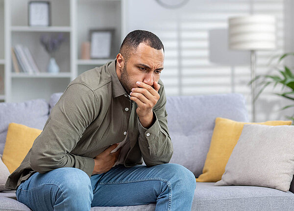 Übelkeit, Erbrechen, Sodbrennen, Durchfall, Verstopfung Ein Mann sitzt vornübergebeugt auf dem Sofa. Er hält sich eine Hand vor den Mund, eine auf den Bauch.