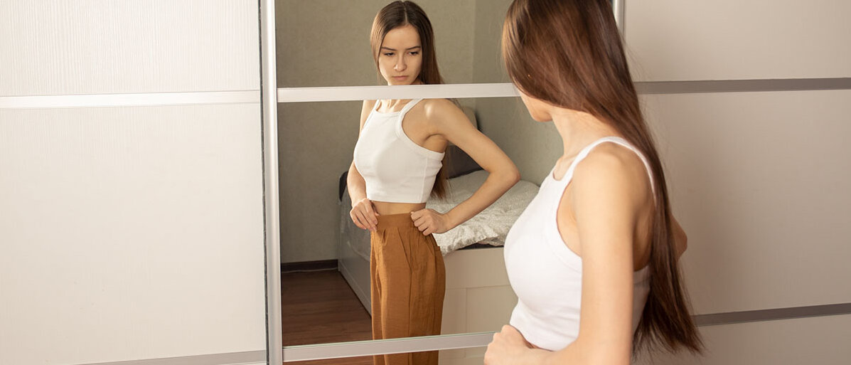 Eine junge Frau steht vor dem Spiegel, betrachtet sich kritisch von der Seite und fasst an ihren Hosenbund. Sie ist sehr dünn.