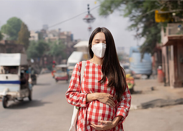 Eine schwangere Frau steht am Straßenrand einer thailändischen Stadt. Sie hat die Hände um ihren dicken Bauch gelegt und trägt eine FFP2-Maske. Im Hintergrund fahren viele kleine Lieferwägen, die Stadt liegt im Dunst.