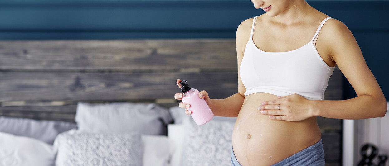 Schwangere Frau mit Cremeflasche in der Hand cremt sich im Schlafzimmer den Bauch ein.