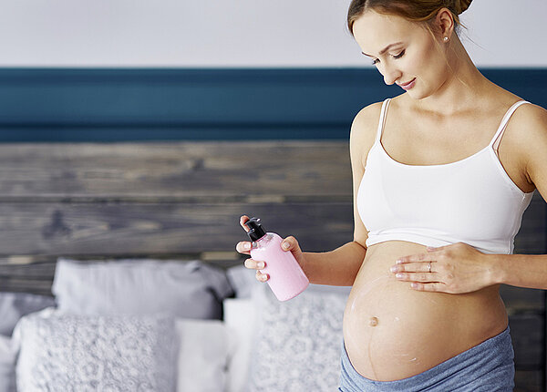 Schwangere Frau mit Cremeflasche in der Hand cremt sich im Schlafzimmer den Bauch ein.