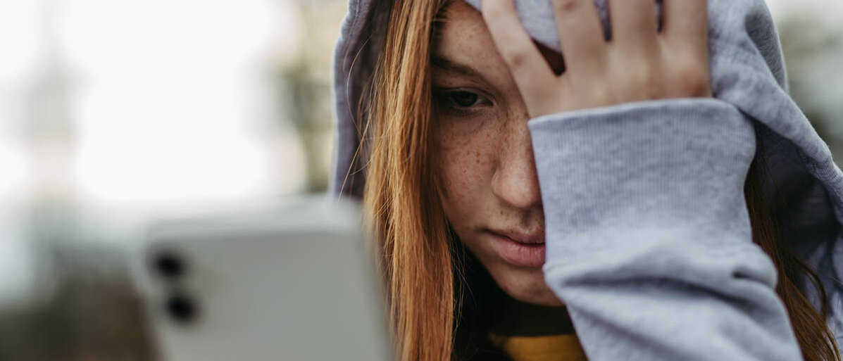 Eine Jugendliche schaut auf ihr Smartphone, stützt die Stirn in die Hand und schaut traurig.