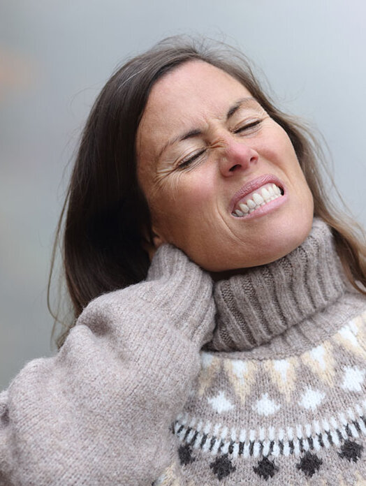 : Eine Frau im Winterpulli bei grauem Wetter greift sich in den Nacken und verzieht vor Schmerzen das Gesicht.