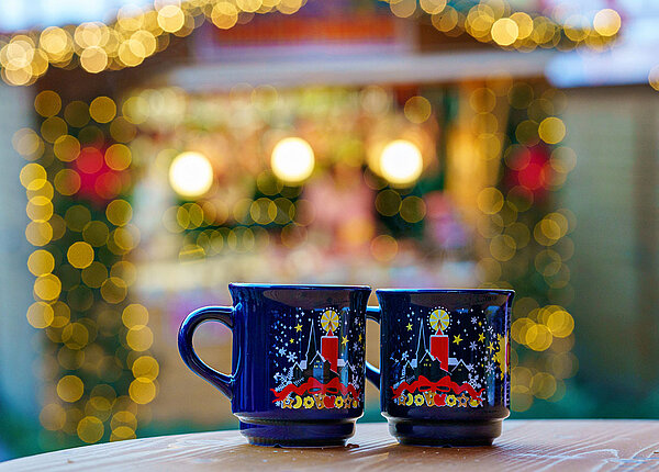 im Vordergrund zwei blaue Tassen mit Glühwein, im Hintergrund ein verschwommener Weihnachtsmarktstand