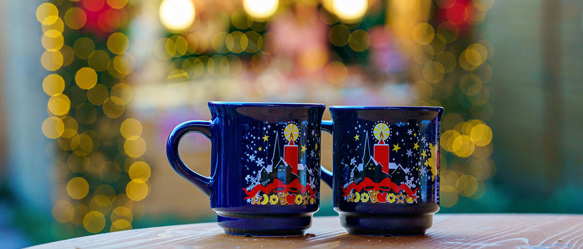 im Vordergrund zwei blaue Tassen mit Glühwein, im Hintergrund ein verschwommener Weihnachtsmarktstand