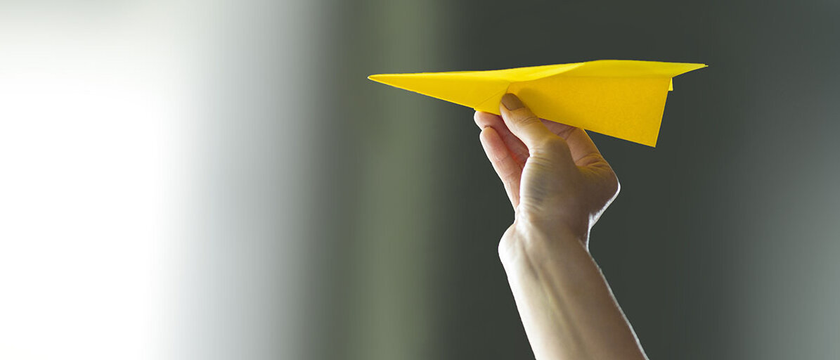 DIE PTA-Newsletter Jemand hält einen gelben Papierflieger.