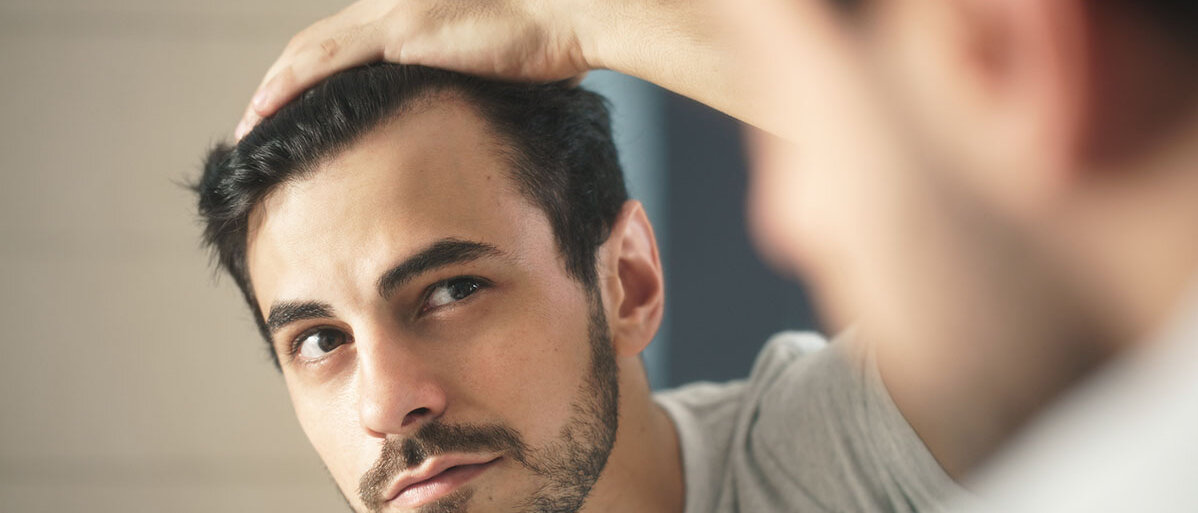 Mann schaut sich im Spiegel seine Haare an und hat eine Hand am Kopf