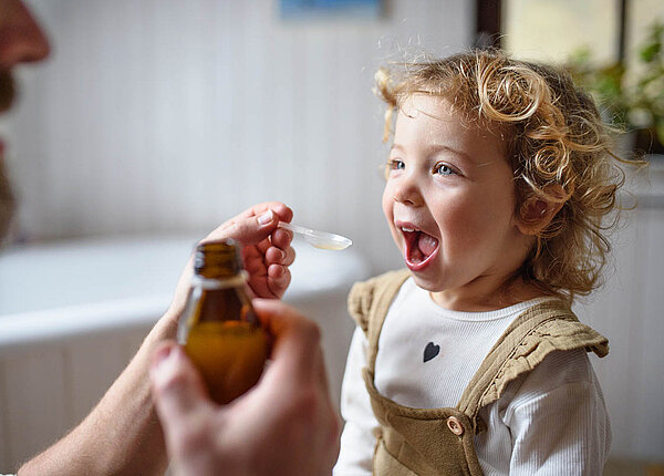 Suspensionen herstellen in der Rezeptur: Praxiswissen und häufige Fehler Ein Mann gibt einem Kleinkind mit einem Dosierlöffel eine Suspension, es hat den Mund weit offen. In der anderen Hand hält der Mann die Braunglasflasche.