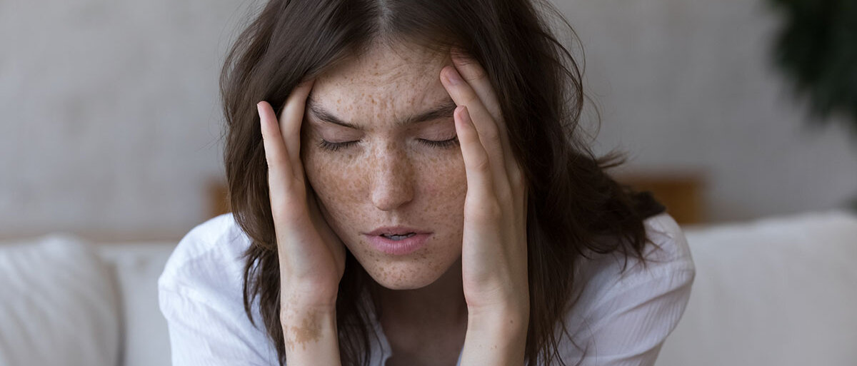 Eine sommersprossige Frau mit dunklen Haaren sitzt auf dem Sofa, hat die Arme auf die Knie gestützt und hält sich die Hände an die Schläfen. Sie hat die Augen geschlossen und das Gesicht verzerrt.