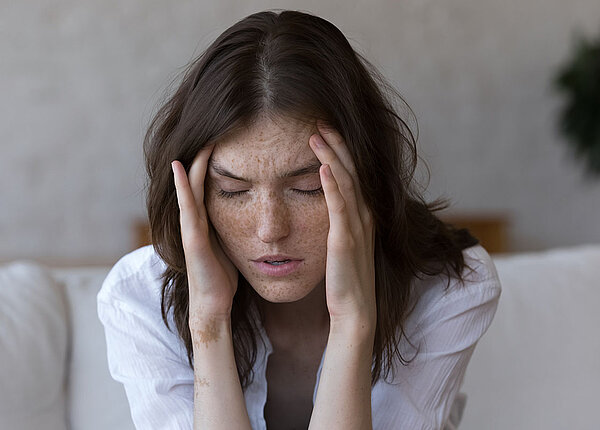 Eine sommersprossige Frau mit dunklen Haaren sitzt auf dem Sofa, hat die Arme auf die Knie gestützt und hält sich die Hände an die Schläfen. Sie hat die Augen geschlossen und das Gesicht verzerrt.