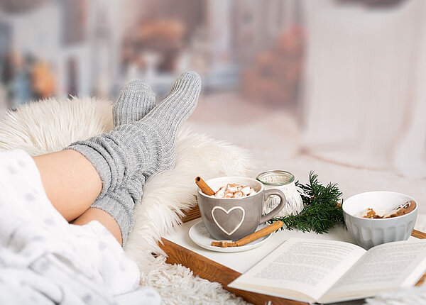 Jemand hat auf einem Fell die Füße hochgelegt, Kuschelsocken an, und daneben steht ein Tablett mit einem heißen Kakao mit Marshmallows, einem aufgeschlagenen Buch, Plätzchen und einer Kerze.