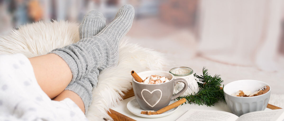 Jemand hat auf einem Fell die Füße hochgelegt, Kuschelsocken an, und daneben steht ein Tablett mit einem heißen Kakao mit Marshmallows, einem aufgeschlagenen Buch, Plätzchen und einer Kerze.