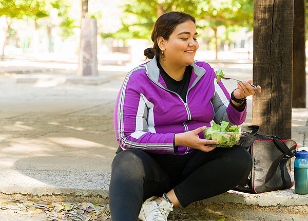 Eine Frau in Sportkleidung sitzt auf einer Stufe und isst genüsslich einen Salat.