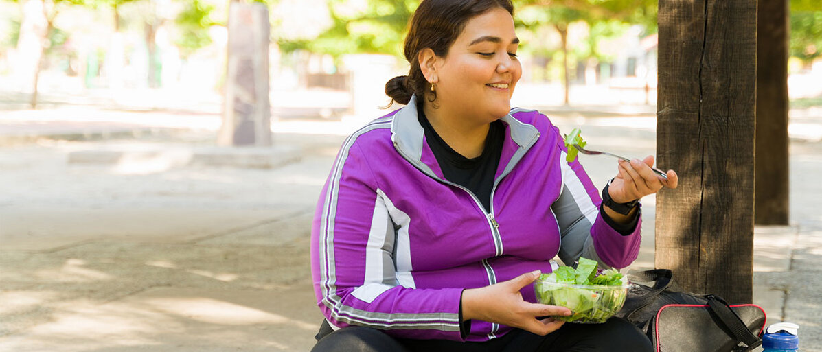 Eine Frau in Sportkleidung sitzt auf einer Stufe und isst genüsslich einen Salat.