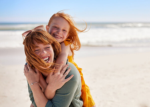 Mädchen umarmt ihre Mutter von hinten und beide lächeln. Beide sind am Strand