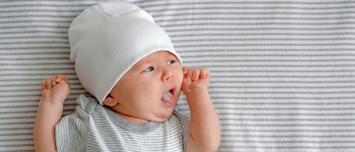 Baby mit weißer Muetze und STrampler liegt auf einer gestreiften Decke und fährt sich mit der Zunge über die Lippen