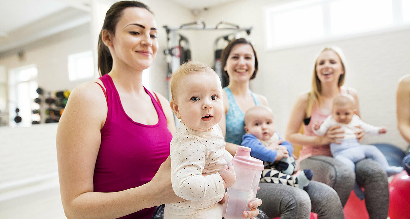 Mehrere jüngere Mütter sitzen in einem Rückbildungskurs auf Gymnastikbällen und haben dabei ihre Babys auf dem Schoß sitzen.