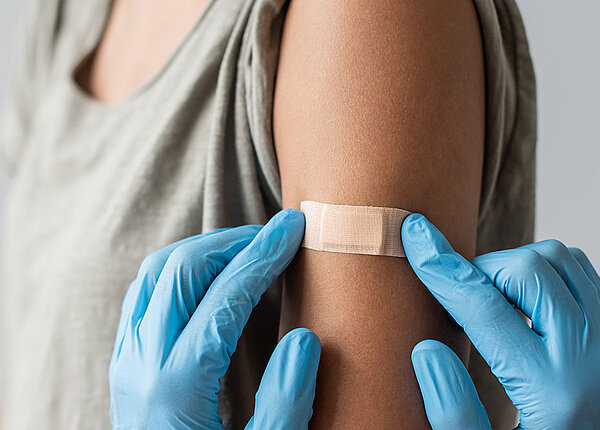  Medizinisches Pesonal mit blauen Handschuhen klebt einer Frau, die den Ärmel ihres T-Shirts hochgestreift hat, ein braunes Pflaster auf den Arm.