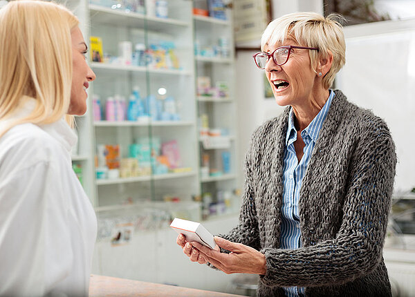 Eine Apothekenkundin hält ein Arzneimittelpäckchen in der Hand und regt sich auf. Die PTA oder Apothekerin lächelt sie ruhig an. 