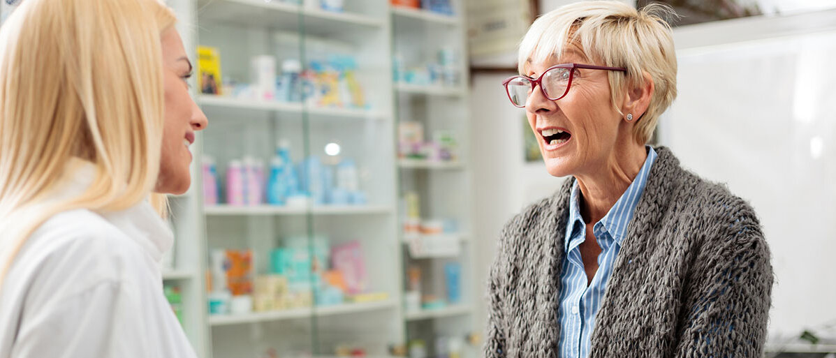 Eine Apothekenkundin hält ein Arzneimittelpäckchen in der Hand und regt sich auf. Die PTA oder Apothekerin lächelt sie ruhig an. 