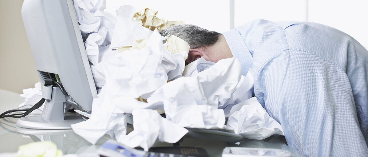 Tipps um Stress am Arbeitsplatz vorzubeugen Mann liegt mit seinem Kopf auf seinem Laptop in einem Meer aus zerknĂŒlltem Papier