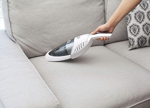 Handstaubsauger Frau reinigt graues Sofa mit Handstaubsauger
