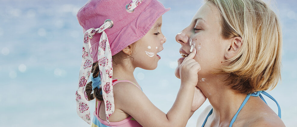 Mutter und Tochter in Badekleidung, Mädchen trägt einen rosa Hut und schmiert das Gesicht ihrer Mutter mit Sonnencreme ein