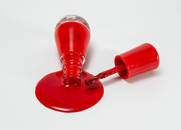 Nagellack richtig entsorgen Roter Nagellack aus einer Glasflasche, der ausgelaufen ist. Nebendran liegt der Pinsel.