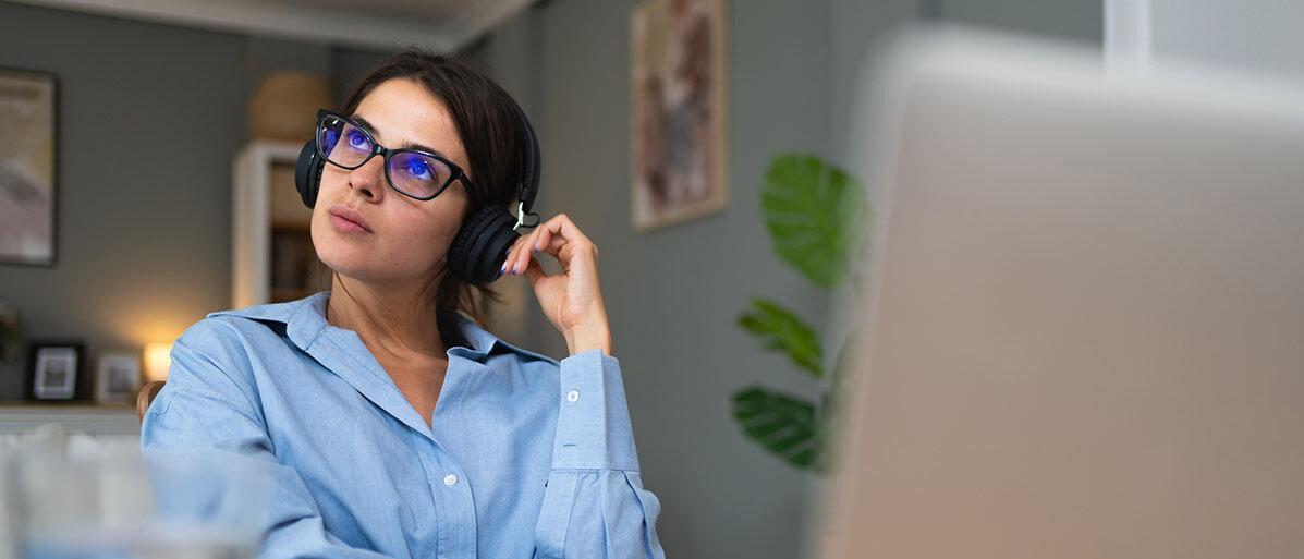 Junge Frau mit Brille sitzt am PC, hat Kopfhörer auf uns schaut verträumt in die Luft