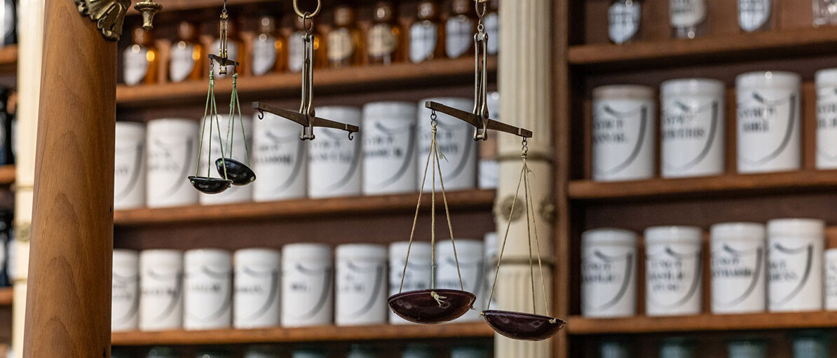 Hilfsstoffe in der Rezeptur: Feuchthaltemittel, Süßungsmittel, Aromastoffe Holzregal einer alten Apothekenrezeptur mit vielen hübschen Standgefäßen. An einer verzierten Halterung hängen drei verschiedene Balkenwaagen.