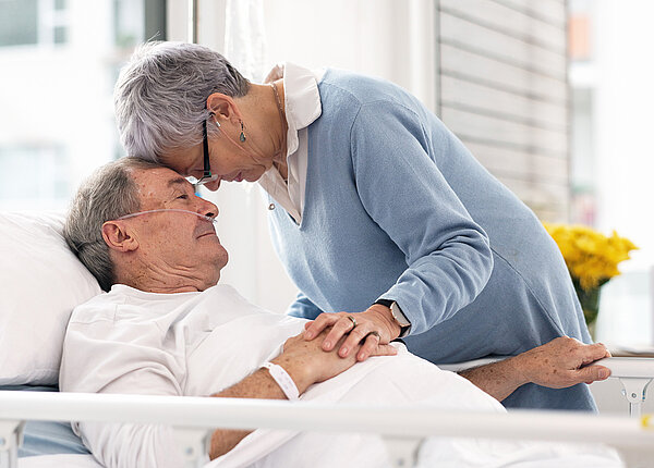 Ältere Frau mit kurzen, grauen Haaren tröstet älteren Mann im Klinikbett. 