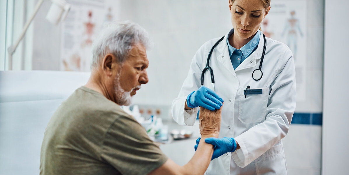 : Ein älterer Mann sitzt auf einer Untersuchungsliege. Eine Ärztin hält die Finger seiner rechten Hand sowie den Unterarm und biegt die Hand nach oben. Im Hintergrund hängen anatomische Zeichnungen.