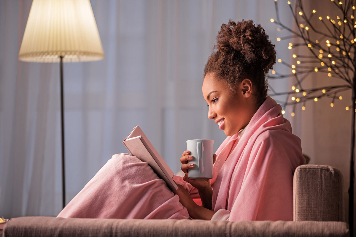Eine junge Frau hat sich auf dem Sofa in eine Decke gewickelt, trinkt aus einer Tasse und liest ein Buch.