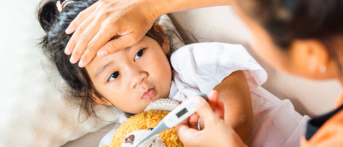 Mutter hat ihre Hand auf der Stirn ihres Kindes, dass vor ihr auf der Couch liegt. Sie hat ein Fieberthermometer in der Hand.