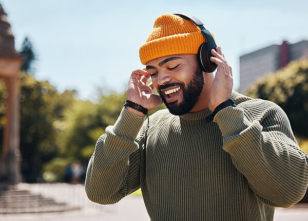 Gut gelaunter, afro-deutscher Mann mit olivgrünem Pulli und orangefarbener Mütze hört Musik über Bluetooth-Kopfhörer 