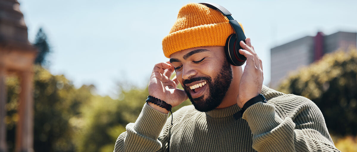 Gut gelaunter, afro-deutscher Mann mit olivgrünem Pulli und orangefarbener Mütze hört Musik über Bluetooth-Kopfhörer 