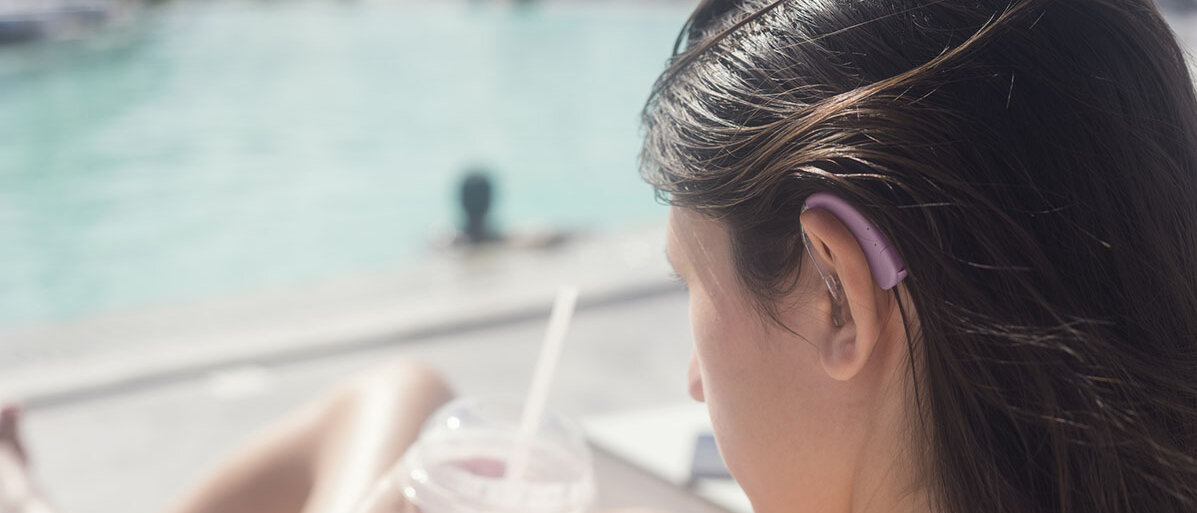 Junge Frau sitzt am Pool mit einem Getränkebecher in der hand und trägt ein lilafarbenes Hörgerät