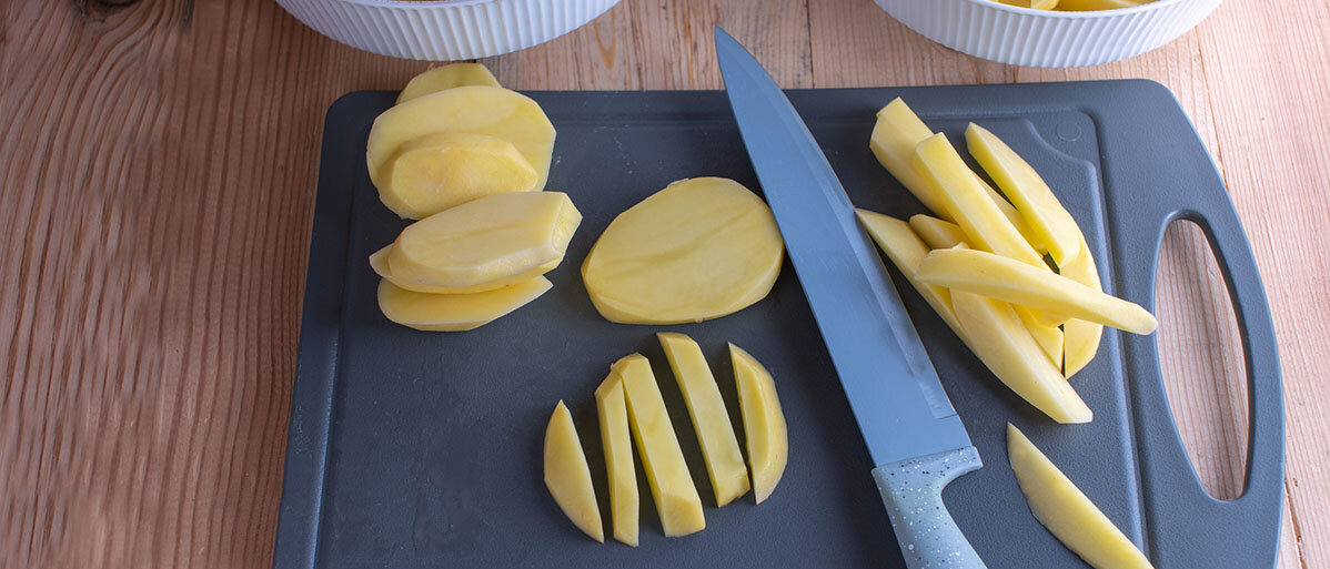 Kartoffelscheiben und bereits geschnittene Kartoffelstreifen liegen auf einem grauen Brettchen. In der Mitte liegt zudem ein Schneidemesser