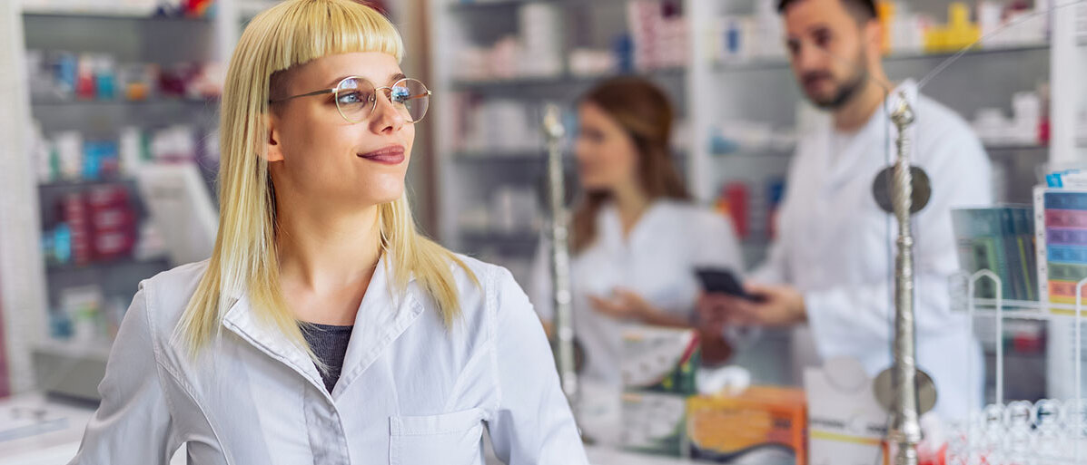 Szene in einer Apotheke: Eine PTA im Vordergrund, ein Apotheker und eine Apothekerin im Hintergrund 