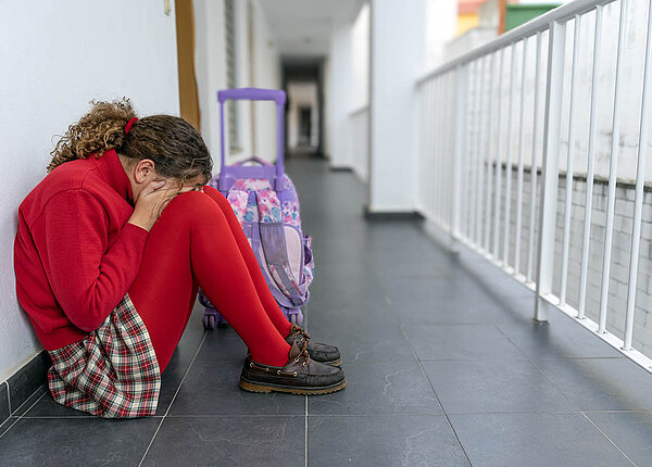 Regelschmerzen von Teenagern vorbeugen und sie behandeln: Welche Medikamente eignen sich bei Jugendlichen mit Periodenschmerzen? Eine Schülerin sitzt allein im Flur der Schule, Beine angezogen, Gesicht in den Händen verborgen.