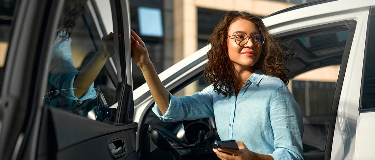 PTA-Beruf: Arbeitsbereiche außerhalb der Apotheke Eine junge Frau steigt aus der Fahrertür eines Autos. Sie trägt eine Bluse, hat ihr Smartphone in der Hand und lächelt.