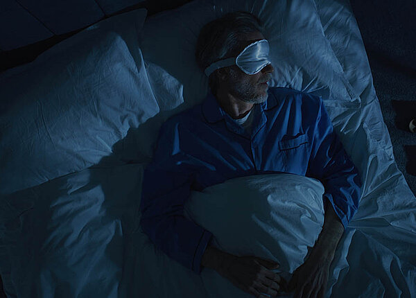 ME/CFS: Behandlung des chronischen Erschöpfungssyndroms dank neuem Biomarker? Ein Mann liegt mit Schlafbrille im dunklen Schlafzimmer im Bett.